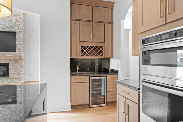 kitchen featuring light wood-type flooring, dark stone countertops, double oven, beverage cooler, and backsplash