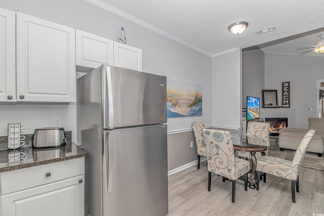 kitchen with a warm lit fireplace, light wood-style flooring, white cabinets, freestanding refrigerator, and dark stone counters