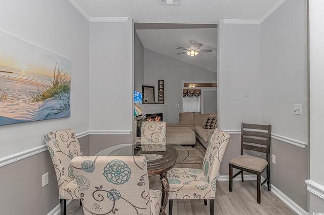 dining area featuring a warm lit fireplace, wood finished floors, a ceiling fan, vaulted ceiling, and ornamental molding