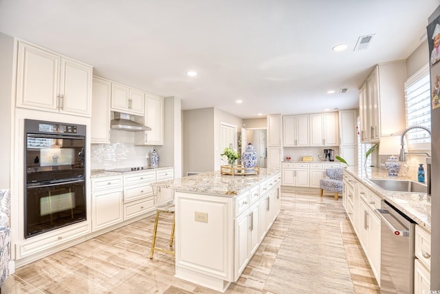 kitchen with a kitchen island, sink, light stone counters, black appliances, and wall chimney range hood