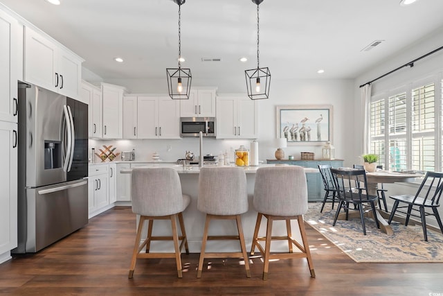 kitchen with appliances with stainless steel finishes, white cabinets, a center island with sink, dark hardwood / wood-style flooring, and decorative light fixtures