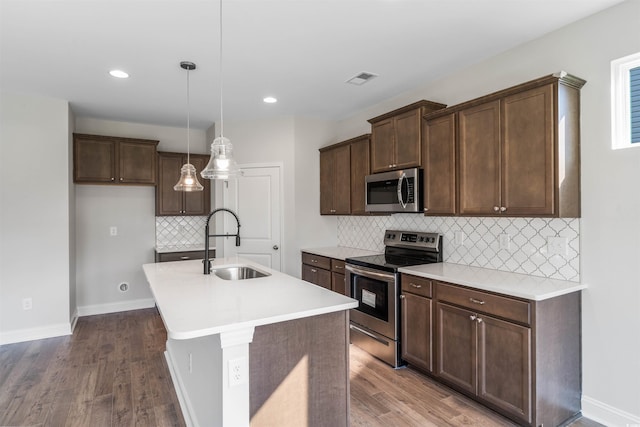 kitchen featuring wood finished floors, decorative backsplash, appliances with stainless steel finishes, a sink, and a center island with sink