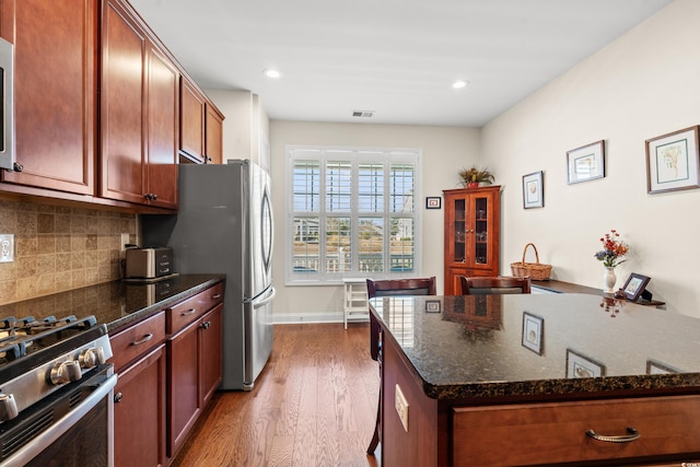 kitchen featuring appliances with stainless steel finishes, dark hardwood / wood-style floors, a kitchen island, decorative backsplash, and dark stone counters