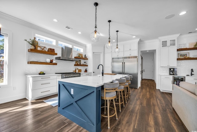 kitchen featuring white cabinets, stainless steel built in fridge, a center island with sink, and wall chimney range hood