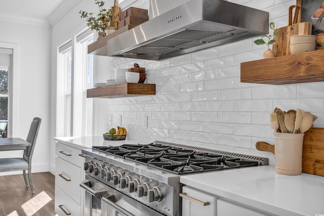 kitchen featuring wall chimney range hood, ornamental molding, high end stove, white cabinets, and decorative backsplash