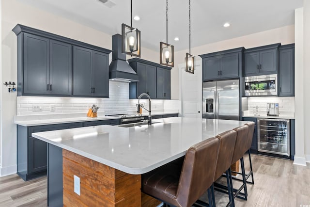 kitchen featuring wine cooler, premium range hood, appliances with stainless steel finishes, pendant lighting, and a kitchen island with sink