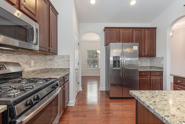 kitchen with dark wood-type flooring, light stone counters, decorative backsplash, arched walkways, and stainless steel appliances