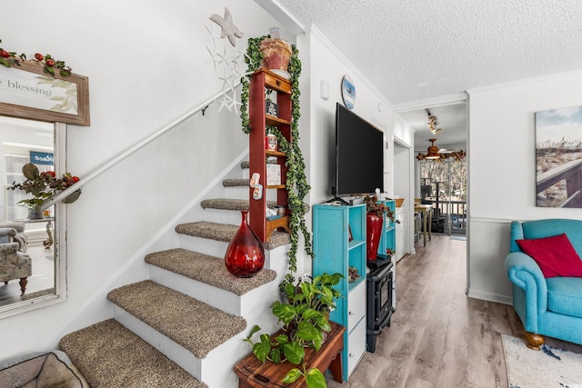 staircase with ceiling fan, wood-type flooring, ornamental molding, and a textured ceiling