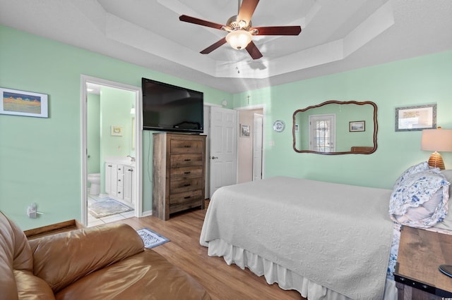 bedroom with a raised ceiling, ensuite bathroom, ceiling fan, and light wood-type flooring