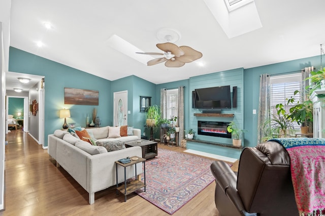 living area featuring vaulted ceiling with skylight, ceiling fan, wood finished floors, and a glass covered fireplace