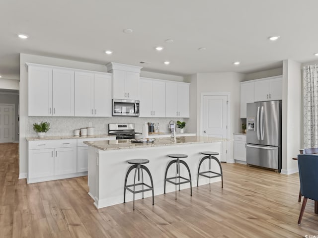 kitchen with stainless steel appliances, white cabinetry, light stone countertops, and a kitchen island with sink