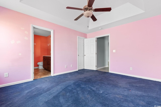 unfurnished bedroom featuring ceiling fan, ensuite bath, a raised ceiling, and dark colored carpet