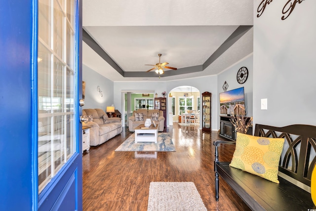 living area featuring arched walkways, a raised ceiling, dark wood-style floors, ceiling fan, and ornamental molding