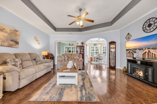 living area with a tray ceiling, arched walkways, dark wood-type flooring, a ceiling fan, and a warm lit fireplace