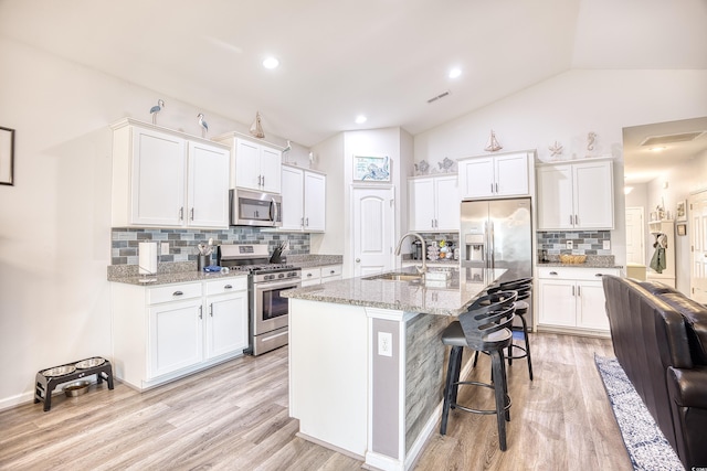 kitchen with sink, a kitchen island with sink, stainless steel appliances, light stone countertops, and white cabinets