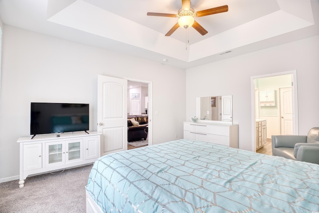 carpeted bedroom with a raised ceiling, connected bathroom, and ceiling fan