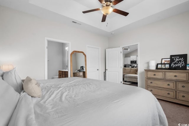 bedroom featuring carpet floors, a tray ceiling, visible vents, ensuite bathroom, and a ceiling fan