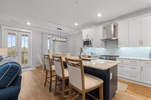 kitchen featuring wall chimney exhaust hood, a center island with sink, stainless steel microwave, ornamental molding, and white cabinets