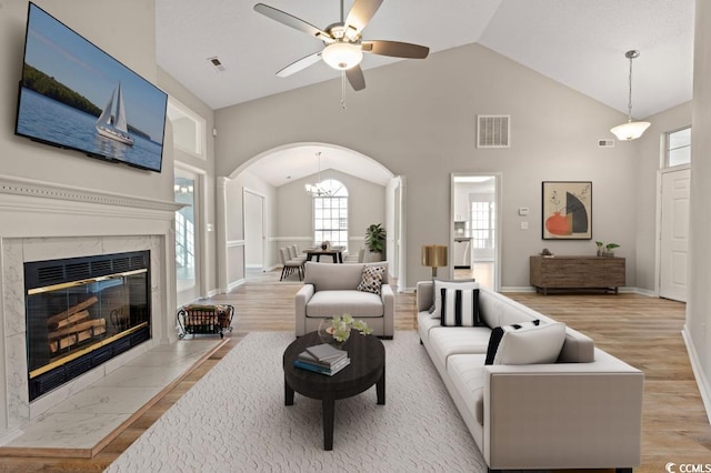 living room featuring high vaulted ceiling, ceiling fan with notable chandelier, a high end fireplace, and light hardwood / wood-style floors
