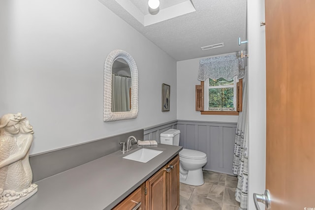 full bath with a textured ceiling, toilet, vanity, visible vents, and wainscoting