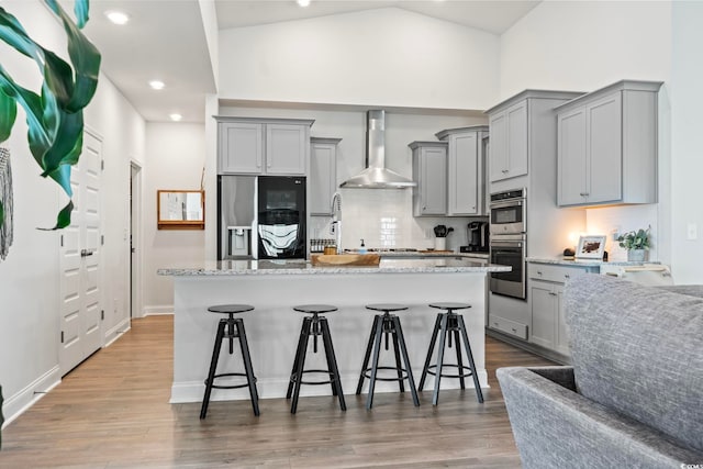 kitchen with wall chimney range hood, stainless steel appliances, a breakfast bar area, and a kitchen island with sink