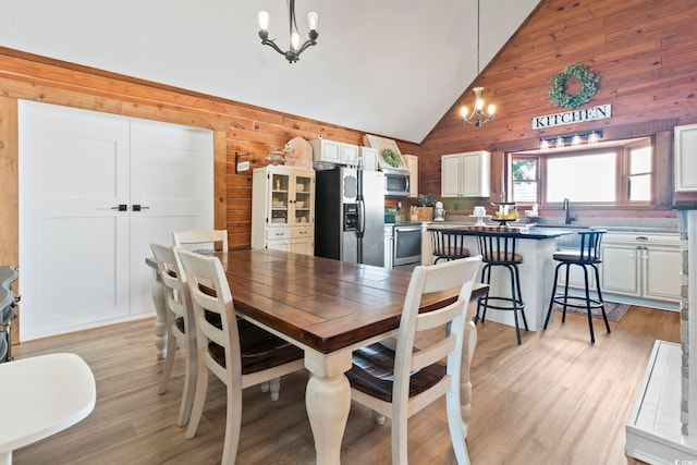 dining space featuring a notable chandelier, light hardwood / wood-style floors, sink, high vaulted ceiling, and wooden walls