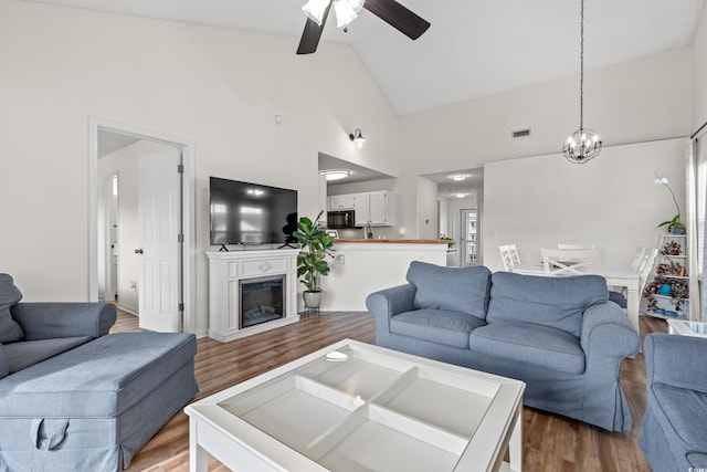 living room with visible vents, a glass covered fireplace, wood finished floors, high vaulted ceiling, and ceiling fan with notable chandelier