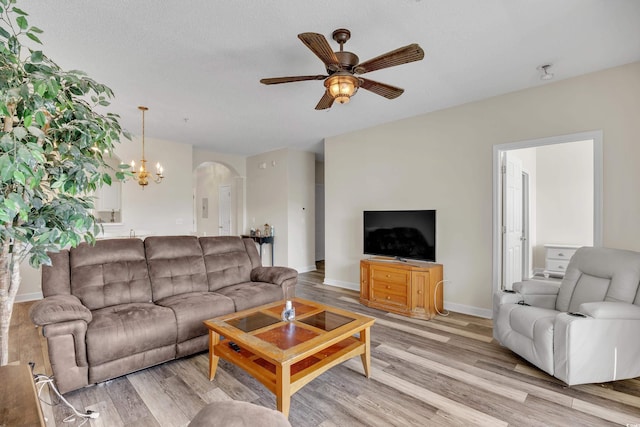 living room featuring light wood-style flooring, baseboards, arched walkways, a textured ceiling, and ceiling fan with notable chandelier