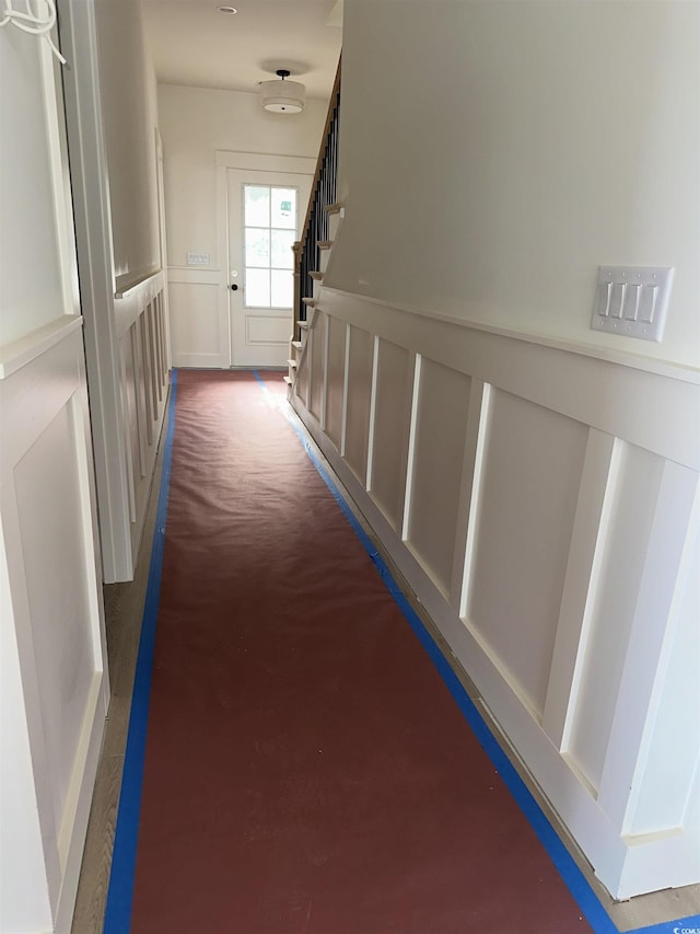 hallway featuring a wainscoted wall, stairs, a decorative wall, and dark colored carpet