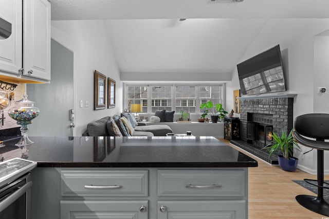 kitchen with lofted ceiling, light wood finished floors, a brick fireplace, and dark countertops