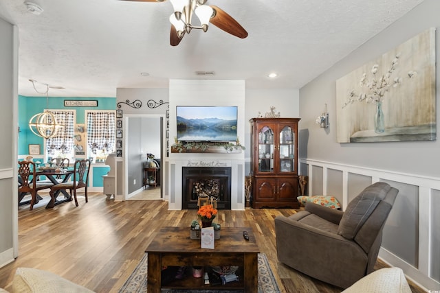 living room featuring a fireplace, visible vents, a decorative wall, wood finished floors, and ceiling fan with notable chandelier