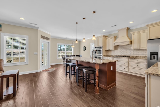 kitchen featuring appliances with stainless steel finishes, light stone counters, decorative light fixtures, cream cabinets, and custom exhaust hood