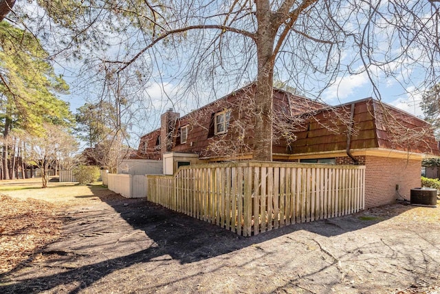 view of side of home with a fenced front yard, brick siding, a chimney, mansard roof, and central AC