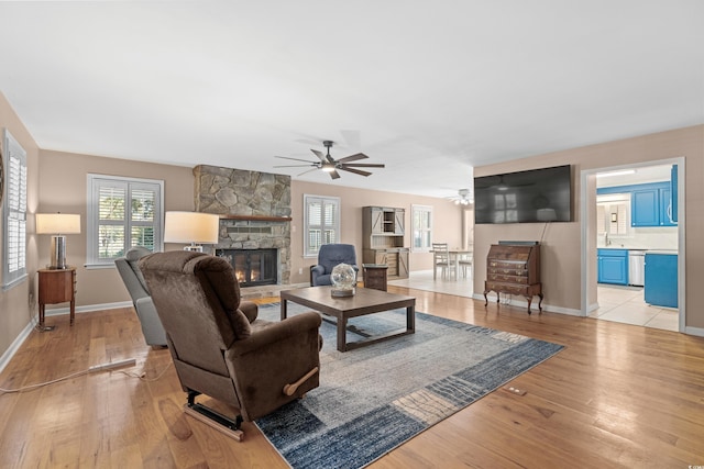 living area featuring light wood-style floors, a stone fireplace, baseboards, and a ceiling fan