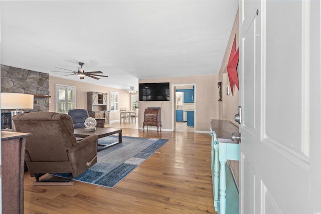 living area with a ceiling fan, baseboards, a stone fireplace, and light wood finished floors