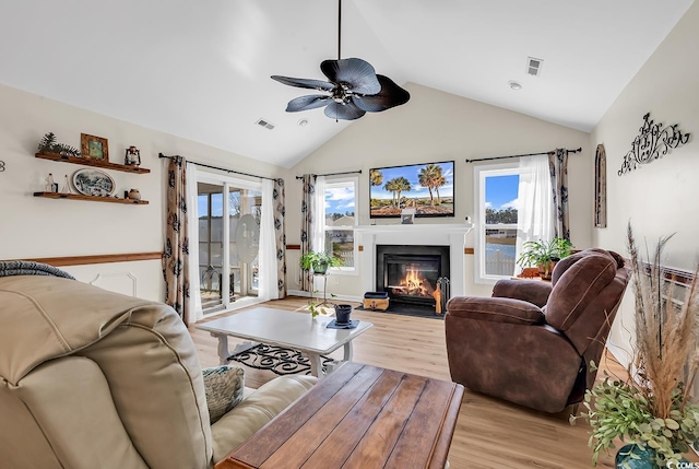 living room featuring light wood finished floors, lofted ceiling, visible vents, a ceiling fan, and a glass covered fireplace