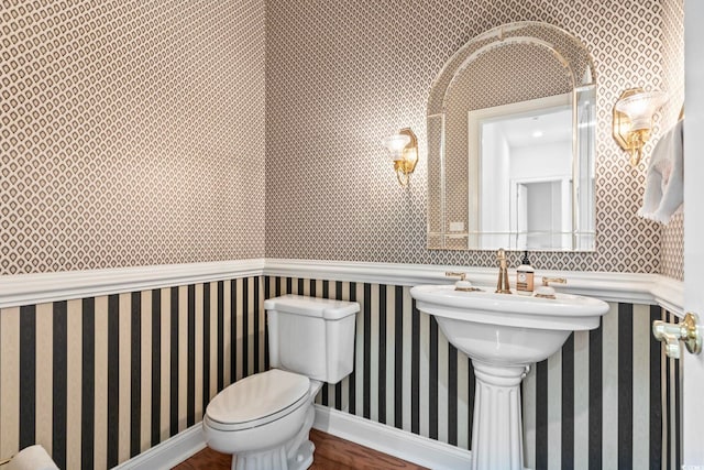 half bath featuring wallpapered walls, baseboards, toilet, a wainscoted wall, and wood finished floors