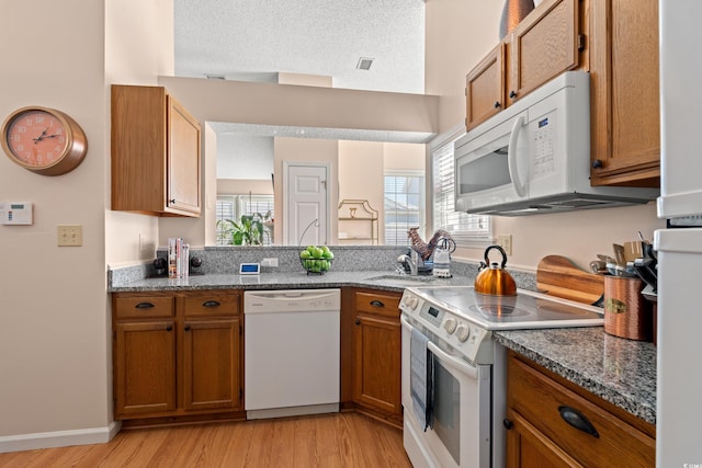 kitchen with a textured ceiling, light wood-style flooring, white appliances, brown cabinets, and dark stone countertops