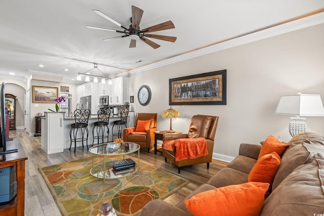 living area with arched walkways, a ceiling fan, baseboards, light wood-type flooring, and crown molding