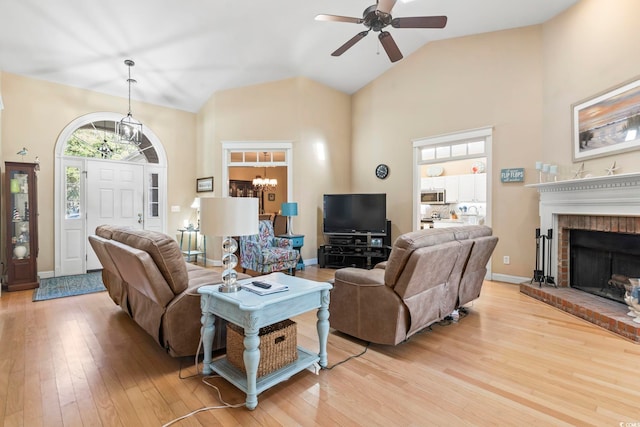 living room with a brick fireplace, baseboards, light wood-style floors, high vaulted ceiling, and ceiling fan with notable chandelier