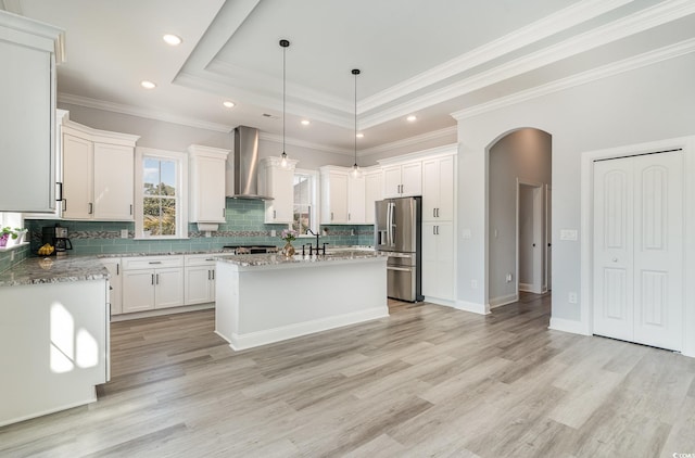 kitchen with stainless steel fridge, arched walkways, a raised ceiling, wall chimney exhaust hood, and a center island