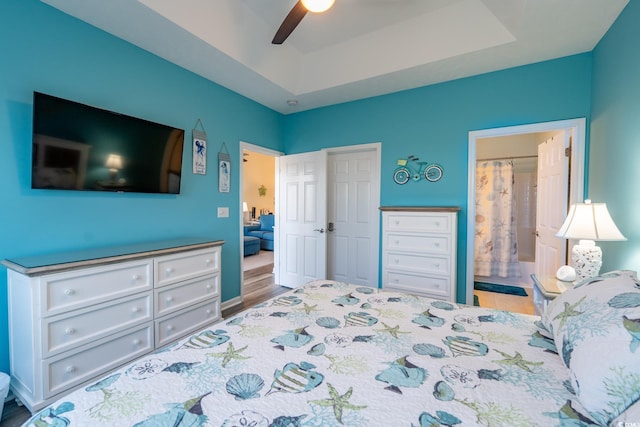 bedroom with wood finished floors, a tray ceiling, connected bathroom, and a ceiling fan