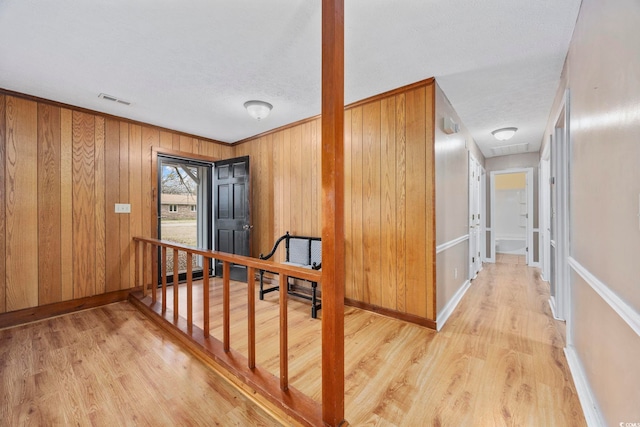 corridor featuring a textured ceiling, wooden walls, visible vents, baseboards, and light wood-style floors