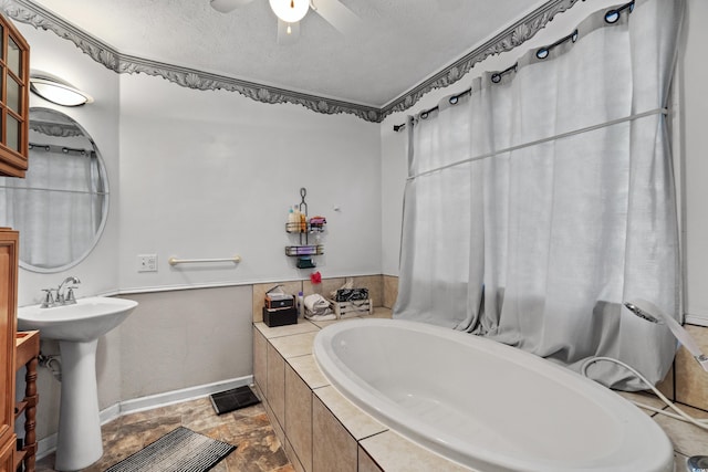 full bath featuring a textured ceiling, ceiling fan, a relaxing tiled tub, baseboards, and stone finish floor