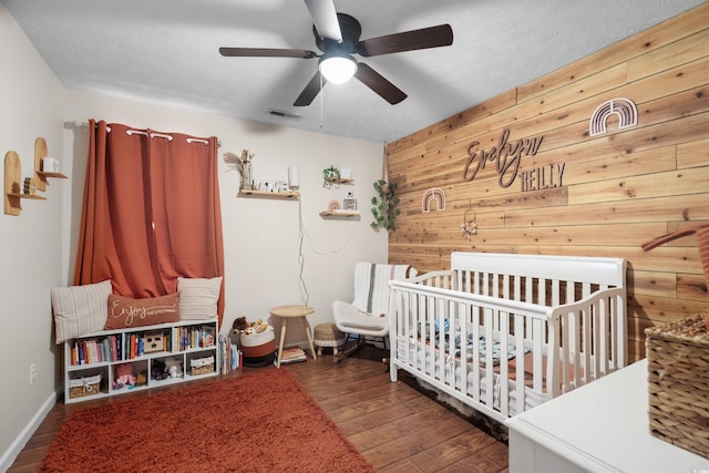 bedroom featuring wooden walls, visible vents, dark wood finished floors, a textured ceiling, and a nursery area