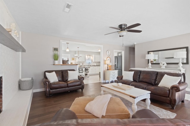 living room with visible vents, a brick fireplace, wood finished floors, baseboards, and ceiling fan with notable chandelier