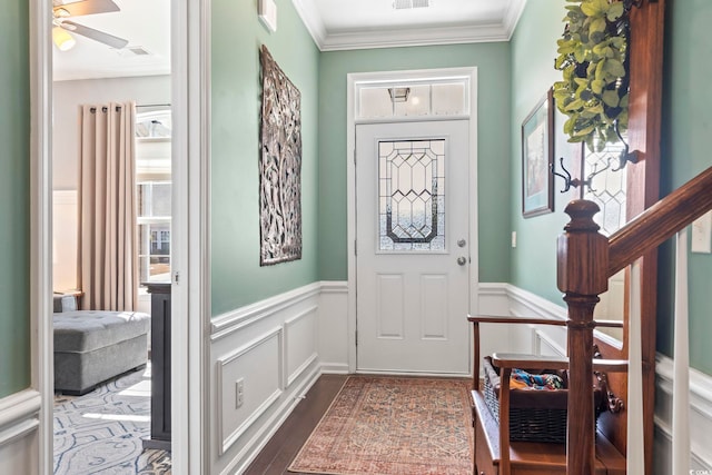 foyer entrance with dark wood finished floors, a ceiling fan, wainscoting, ornamental molding, and stairs