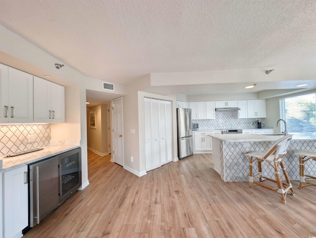 kitchen featuring beverage cooler, white cabinets, appliances with stainless steel finishes, under cabinet range hood, and a kitchen bar