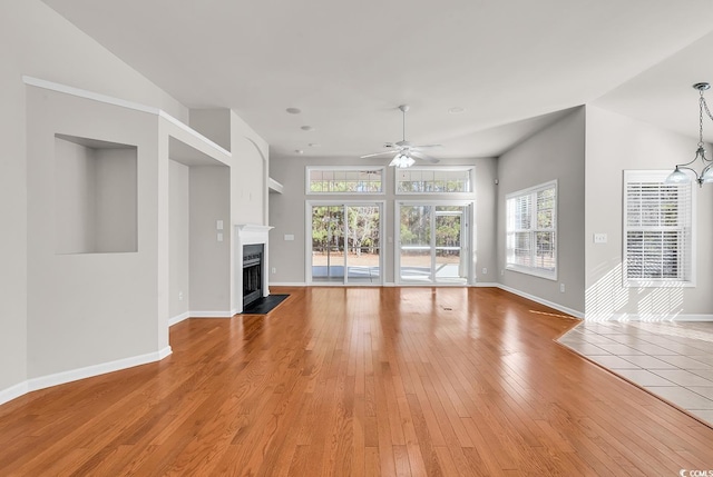 unfurnished living room featuring a fireplace with flush hearth, baseboards, light wood finished floors, and ceiling fan with notable chandelier