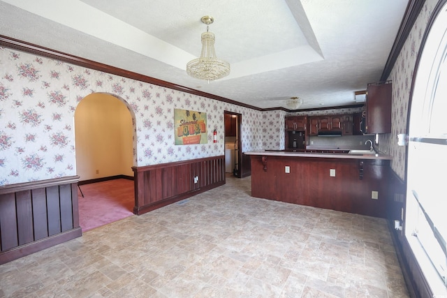 kitchen with arched walkways, a tray ceiling, wainscoting, a textured ceiling, and wallpapered walls